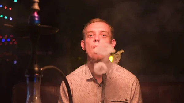 Nuori miesmalli tekee savua hookah-renkaista istuessaan nahkasohvalla. Media. Muotokuva miehestä, jolla on suuret silmät ja joka hengittää savupilviä suustaan.. — kuvapankkivalokuva