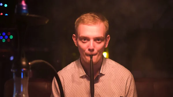 Young blond man with big green eyes smoking shisha in restaurant. Media. Man resting with hookah looking straight to the camera late in the evening. — Stock Photo, Image