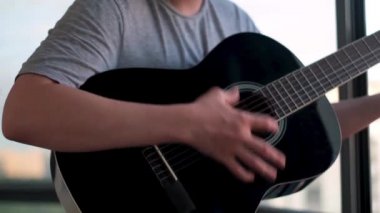 Balkonda pencerelere karşı duran ve gitar çalan bir adam. Kavram. Genç adam evde akustik gitar, sosyal uzaklık ve covid 19 konseptiyle pratik yapıyor..