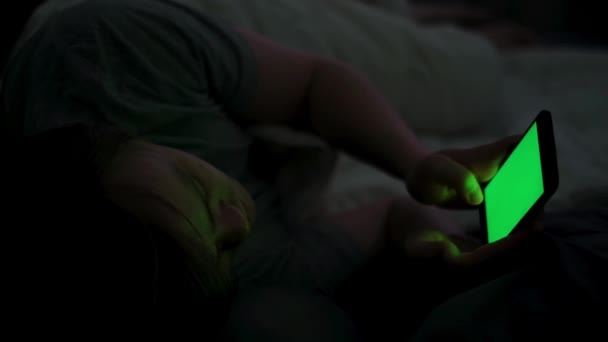 Jovem viciado em smartphone com tela verde na cama à noite. Conceito. Close-up de homem que sofre de insônia usando seu smartphone com chave chroma enquanto deitado em sua cama à noite. — Vídeo de Stock