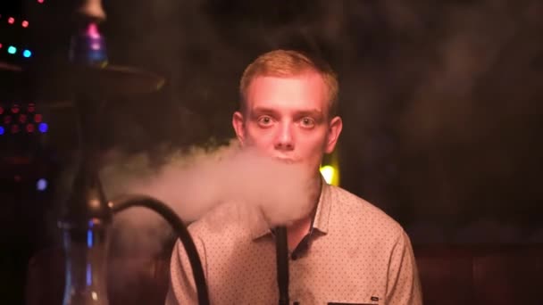Young blond man with big green eyes smoking shisha in restaurant. Media. Man resting with hookah looking straight to the camera late in the evening. — Stock Video