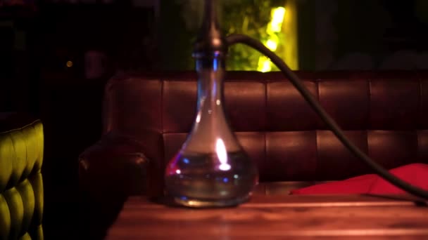 Gros plan du narguilé fait à la main placé sur une table en bois dans un bar narguilé. Les médias. Ambiance chaleureuse et la fiole en verre shisha sur le canapé en cuir et vert néon lampe arrière-plan. — Video