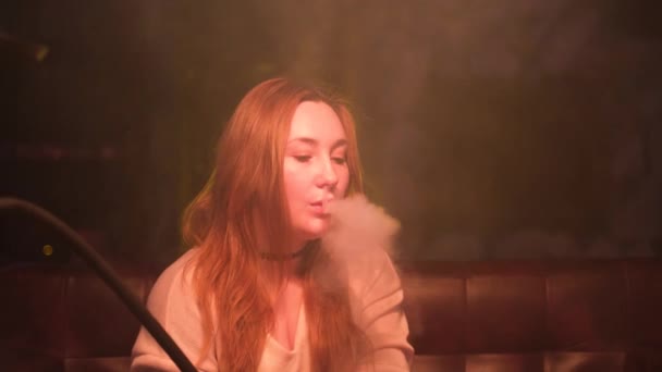 Hermosa chica con el pelo largo rojo y labios sensibles sentados en el sofá y fumar narguile. Medios. Melancólica jovencita fumando shisha, exhalando humo por la nariz y la boca. — Vídeos de Stock