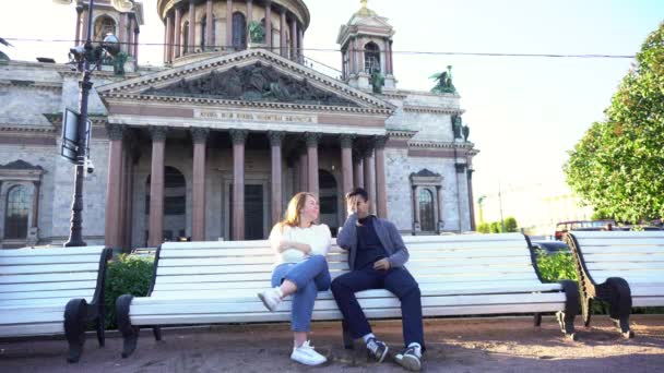 Jeune couple assis sur un banc près de la cathédrale Saint-Isaac à Saint-Pétersbourg, Russie. Les médias. Homme et femme assis ensemble et bavardant comme des amis. — Video