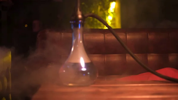 Glazen haakje met glazen schaaltje op tafel in loungebar. De media. Sluiten van shisha flacon in rookwolken op een houten tafel in een hookah cafe op de achtergrond van geen neon lamp. — Stockfoto