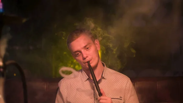 Ein Mann schaut nachdenklich, während er Rauch aus seinem Mund ausatmet. Medien. Schöner kaukasischer Mann in weißem Hemd hält eine Wasserpfeife im Mund und raucht. — Stockfoto