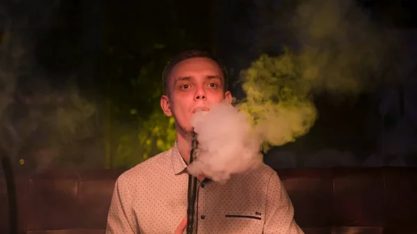 A guy looking pensive while exhales smoke from his mouth. Media. Handsome caucasian man in a white shirt holds a hookah pipe in her mouth and smokes. — Stock Photo, Image