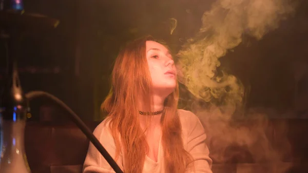 Menina bonita com cabelos longos vermelhos e lábios sensíveis sentados no sofá e fumando narguilé. Mídia. Jovem melancólica fumando shisha, exalando fumaça pelo nariz e boca. — Fotografia de Stock