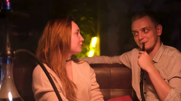 Bărbat şi femeie la o întâlnire la un bar de narghilea fumând şi vorbind. Media. Fumătorul de sex masculin ascultă cu atenție prietena lui în timp ce fumează shisha sub lumina slabă . — Fotografie, imagine de stoc