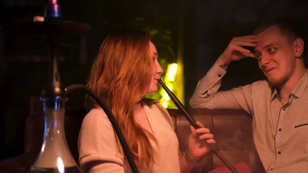 Happy couple interacting in night club while smoking shisha. Media. Young man and woman on a date talking to each other, having fun, girl exhaling clouds of smoke, concept of love. — Stock Photo, Image