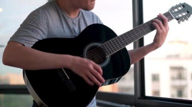 Balkonda pencerelere karşı duran ve gitar çalan bir adam. Kavram. Genç adam evde akustik gitar, sosyal uzaklık ve covid 19 konseptiyle pratik yapıyor..