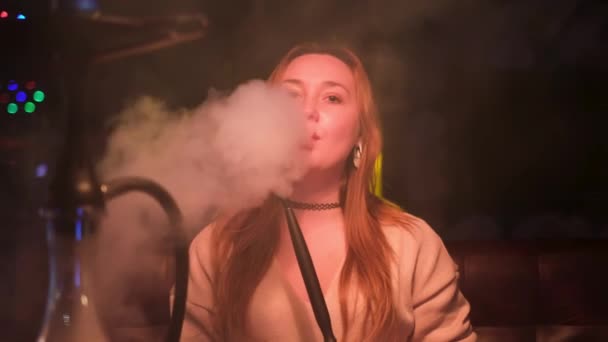 Hermosa mujer joven sexy con maquillaje natural vapeo en discoteca. Medios. Mujer exhalando humo en el bar y mirando directamente a la cámara. — Vídeos de Stock