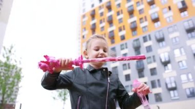 Dişi bir çocuğun köpük üflemesi ve gülmesi. Başla. Sevimli küçük kız, sonbahar günü çok katlı binanın avlusunda eğlenirken sabun köpüğü üflüyor..