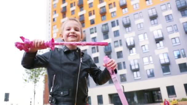 Vista in basso di una bambina che soffia bolle e ride. Azione. Adorabile bambina sta soffiando bolle di sapone nella giornata autunnale mentre si diverte nel cortile dell'edificio a più piani. — Video Stock