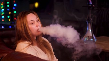 Gece kulübünün karanlık arka planında nargile içen bir kadın. Medya. Artı, sigara içme alışkanlığı olan güzel bir model, boş zaman kavramı ve rahatlama..