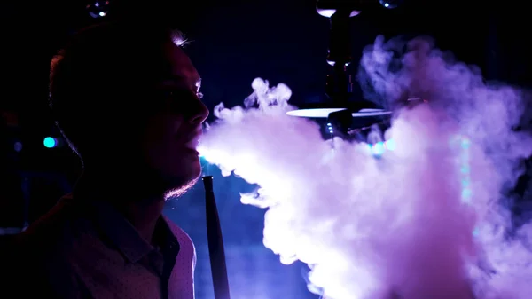 Neon ışıkta erkek siluetinin yan görüntüsü nargile tüttürüyor, gece kulübünde dinleniyor. Medya. Sigara içen adam ağzından dumanlar salıyor.. — Stok fotoğraf