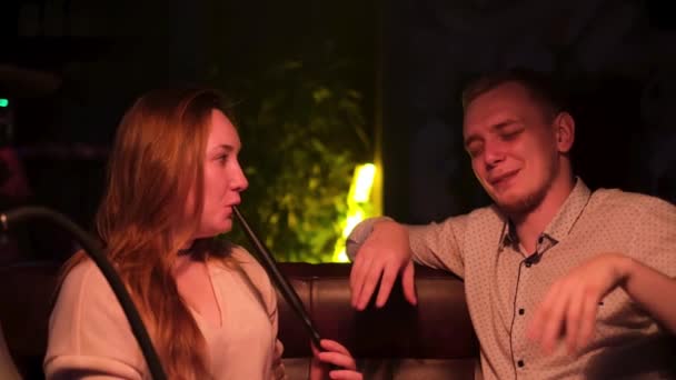 Un couple fumant de la chicha au bar du narguilé. Les médias. Homme et femme assis sur le canapé, mec fait une blague pour faire rire sa copine pendant qu'elle tient un tube de narguilé. — Video