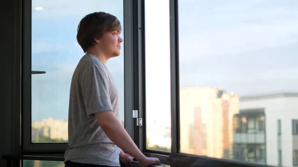 Un homme adulte en t-shirt gris debout contre une fenêtre ouverte sur le balcon et profitant de la vue. Concept. Vue latérale d'un homme heureux regardant dans le nouveau jour par la fenêtre ouverte à la maison. — Photo
