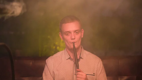 Jeune homme blond fumant narguilé et produisant des anneaux de fumée. Les médias. Fumeurs magiques. jeune homme relaxant avec shisha, soufflant facilement des anneaux de fumée. — Photo