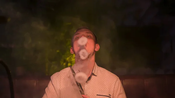 Homem caucasiano exalando uma nuvem de nevoeiro e arrepiante. Mídia. Vista frontal de cara bonito jovem fumando narguilé sozinho e se sentindo deprimido no quarto escuro do salão shisha. — Fotografia de Stock