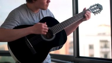 Genç bir adam ya da müzisyen evdeki pencereye karşı gitar çalıyor. Kavram. Siyah akustik gitar tutan ve akor çalan bir adama yaklaş..