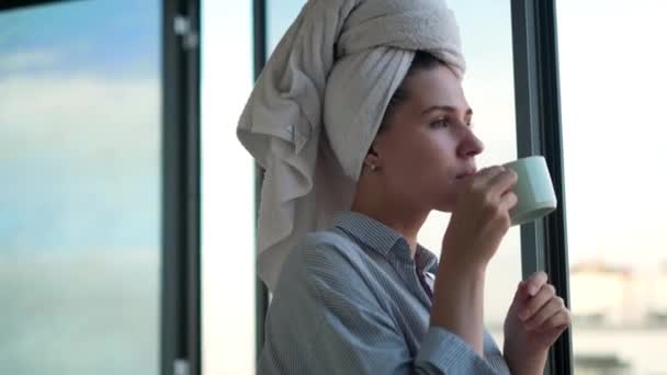 Kobieta z włosami owiniętymi w biały ręcznik, delektująca się poranną kawą lub herbatą na balkonie. Koncepcja. Portret pięknej dziewczyny pijącej gorący napój przy otwartym oknie. — Wideo stockowe
