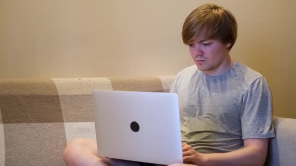 家の服を着たラップトップでソファに座っている若い男に集中しました。コンセプト。家庭で働く真剣なブロンドの男、インターネットと彼のコンピュータを使用して、フリーランスの概念. — ストック動画