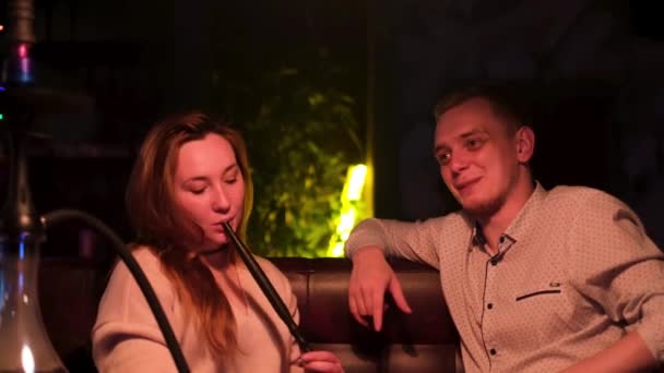 Couple smoking shisha in hookah bar. Media. Man and woman sitting on the couch, guy is making a joke to make her girlfriend laugh while she is holding a hookah tube. — Stock Video