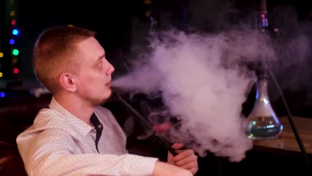 Side view of a man wearing white shirt smoking hookah and exhaling white smoke. Media. Young man sitting on black leather sofa and smoking shisha on the background of night club or bar room. — Stock Video