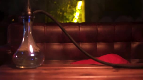 Glass hookah with glass bowl standing on the table in lounge bar. Media. Close up of shisha flask in clouds of smoke on a wooden table in a hookah cafe on the background of geen neon light lamp. — Stock Video