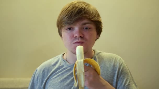 Portret van een volwassen hongerige man die een rijpe banaan met eetlust eet. Concept. Vooraanzicht van een man in grijs t-shirt bijten fruit en kauwen. — Stockvideo