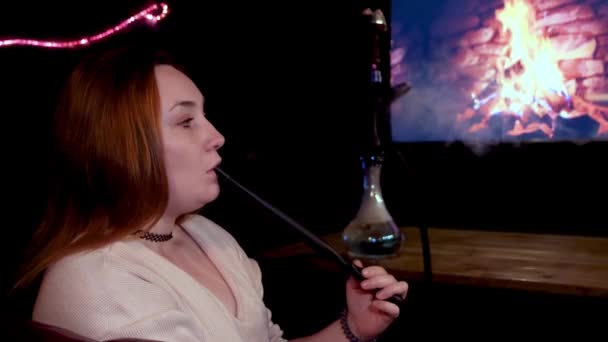 Vista lateral de una chica pelirroja fumando shisha frente a la pantalla del televisor con fuego ardiente. Medios. Joven hermosa mujer con una gargantilla negro alrededor de su cuello fumar narguile en el fondo de la falsificación — Vídeos de Stock