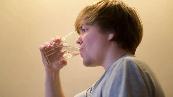 Sidovy över en blond kaukasisk man som dricker klart vatten. Begreppet. Törstig ung kille som håller glas transparent glas vatten och dricka isolerad på gul vägg bakgrund. — Stockfoto