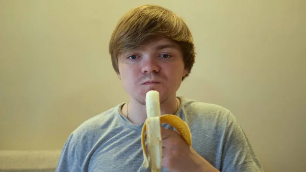 Retrato de um adulto faminto que come uma banana madura com apetite. Conceito. Vista frontal de um cara em camiseta cinza mordendo frutas e mastigar. — Fotografia de Stock