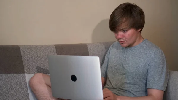 Een lachende aantrekkelijke jongeman gebruikt zijn laptop thuis op de bank in de woonkamer. Concept. Blond man draagt thuis grijs t-shirt tijdens het typen op zijn computer. — Stockfoto