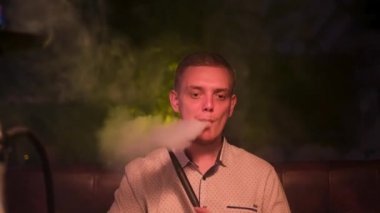 Genç sarışın adam nargile içiyor ve duman halkaları üretiyor. Medya. Sihirli sigara içmek. Genç adam shisha ile dinleniyor, kolayca duman halkaları üflüyor..