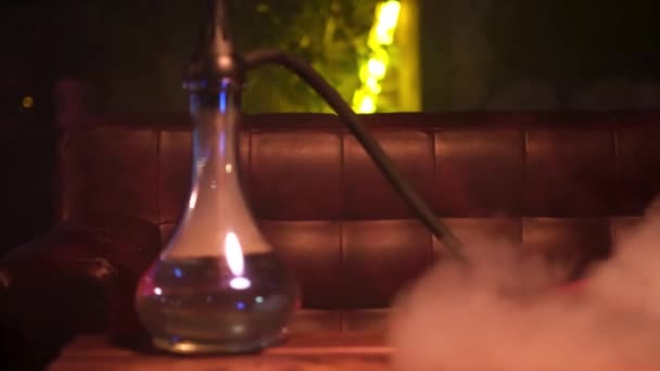 Glass hookah with glass bowl standing on the table in lounge bar. Media. Close up of shisha flask in clouds of smoke on a wooden table in a hookah cafe on the background of geen neon light lamp. — Stock Video