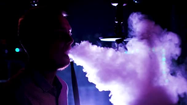 Boční pohled na mužskou siluetu v neonovém světle kouřící hookah, relaxace v nočním klubu. Média. Muž kouřící shishu, vydechující hustý kouř z úst. — Stock video