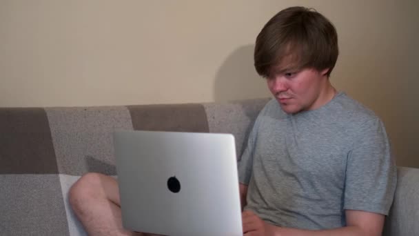 Uśmiechnięty, atrakcyjny młodzieniec używa laptopa, siedząc w domu na kanapie w salonie. Koncepcja. Blondyn w szarej koszuli podczas pisania na komputerze. — Wideo stockowe