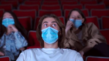 Sinemada film izlerken tıbbi maske takan korkmuş genç adama yaklaş. Medya. Beyaz gömlekli yakışıklı bir adamın portresi korku izlerken şok olmuş görünüyor..