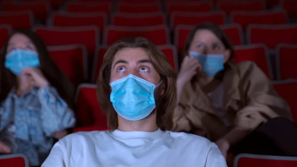 Detailní záběr vyděšeného mladého muže v lékařské masce při sledování filmu v kině. Média. Portrét pohledného muže v bílé košili vypadá šokovaně při sledování hororu. — Stock fotografie