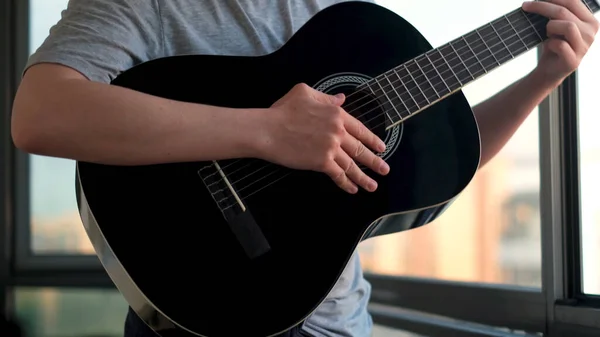 Un homme avec une guitare à la main se tient près de la fenêtre du balcon. Concept. Musicien avec une guitare acoustique noire jouant des chansons à la maison. — Photo