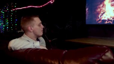 Genç adam şöminenin yanında nargile içerken dinleniyor. Medya. Şöminenin yanında sigara içen bir adam TV ekranında resmedilmiş..