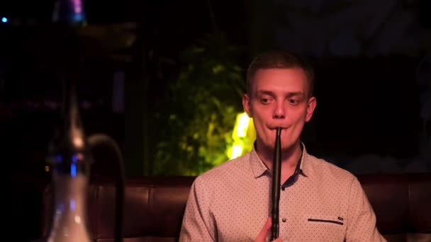 Hombre caucásico exhalando una nube de niebla y enfriándose. Medios. Vista frontal de un joven guapo fumando narguile solo y sintiéndose deprimido en la habitación oscura del salón shisha. — Vídeos de Stock