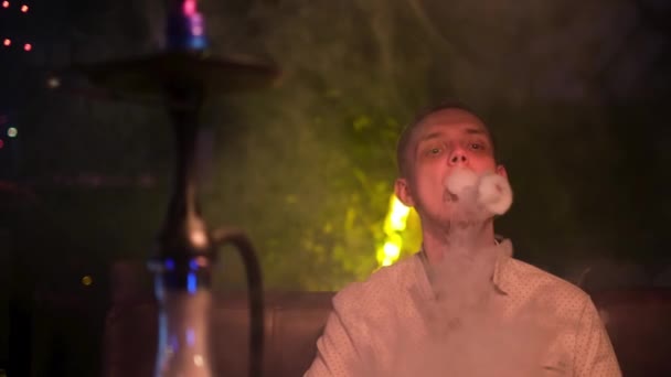 Genç sarışın adam nargile içiyor ve duman halkaları üretiyor. Medya. Sihirli sigara içmek. Genç adam shisha ile dinleniyor, kolayca duman halkaları üflüyor.. — Stok video