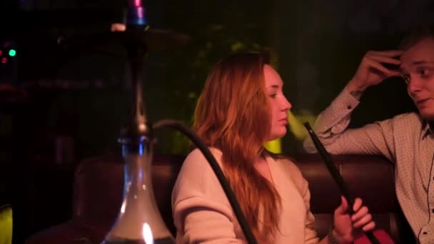 Happy couple interacting in night club while smoking shisha. Media. Young man and woman on a date talking to each other, having fun, girl exhaling clouds of smoke, concept of love. — Stock Video