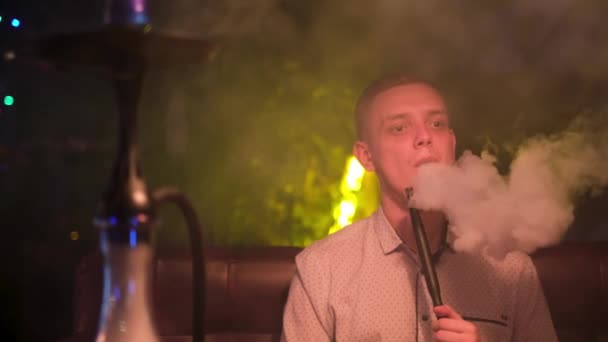 Чоловік, який виглядає пухнастим, вдихає дим з рота. Медіа. Красивий білий чоловік у білій сорочці тримає кальянну трубу в роті і курить . — стокове відео