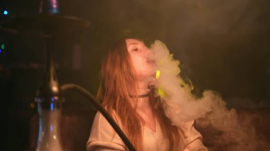 Koyu yakalı ve nargile dumanı doğrudan kameraya üfleyen seksi kızıl saçlı kız. Medya. Güzel bir kadın sigara içerken vakit geçiriyor..