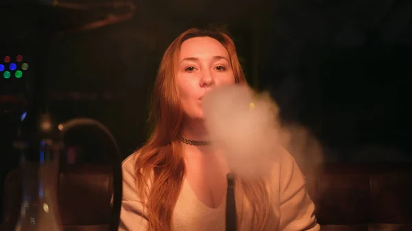 Red haired sexy girl in a lounge bar smoking a hookah dressed in light brown sweater on dark interior background. Media. Beautiful lady looking playfully and blowing clouds of smoke. — Stock Photo, Image