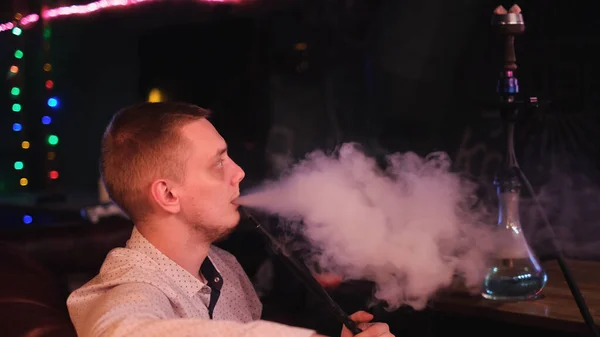 Jongeman is verslaafd aan een hookah. De media. Close-up zijaanzicht van een man in shirt uitademen dikke rook uit zijn mond om donkere achtergrond van cafe interieur. — Stockfoto